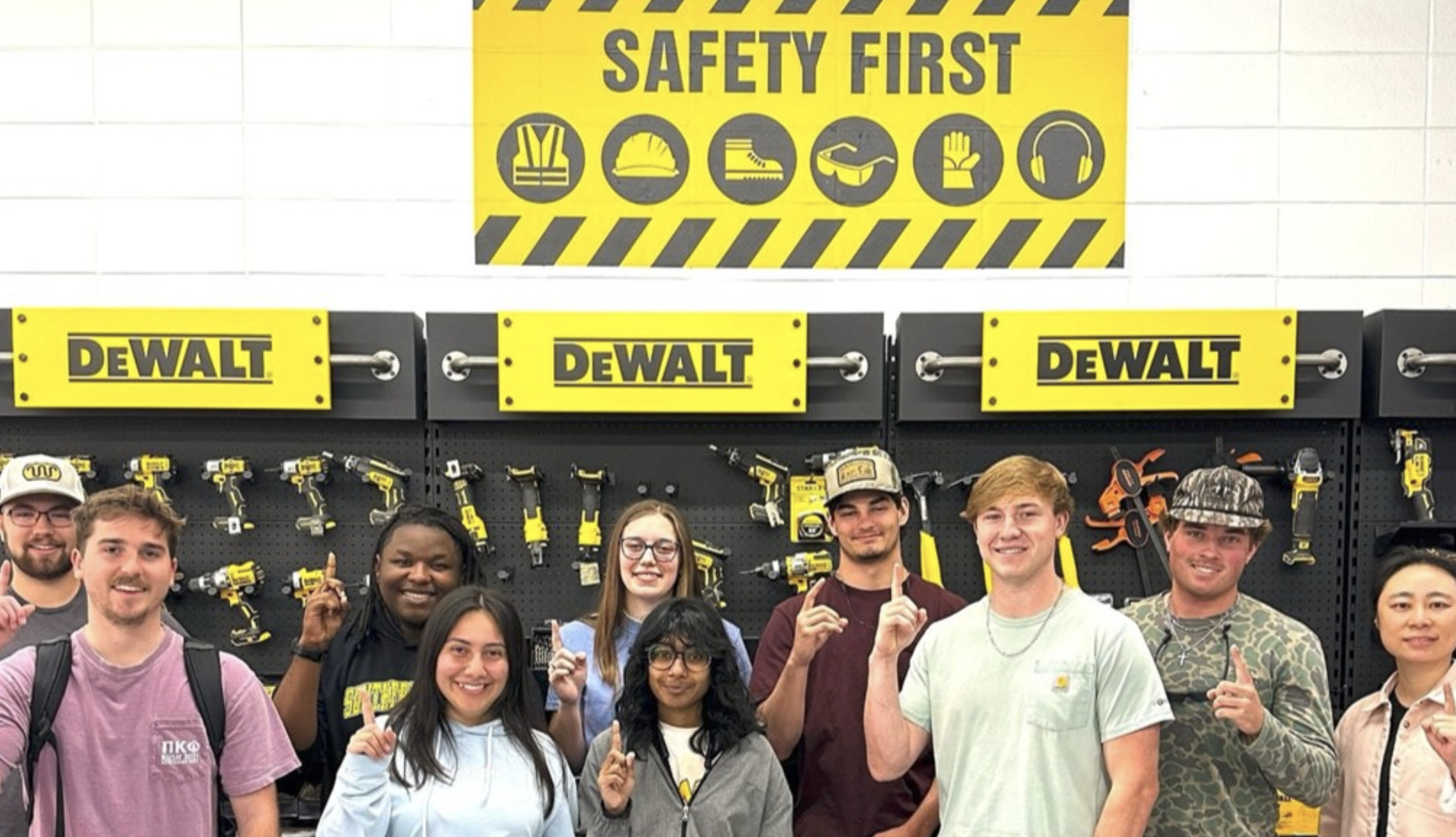 group photo of Dr. Zhang and students in front of a tool wall with yellow signs that read DeWalt and a larger yellow "Safety First" sign